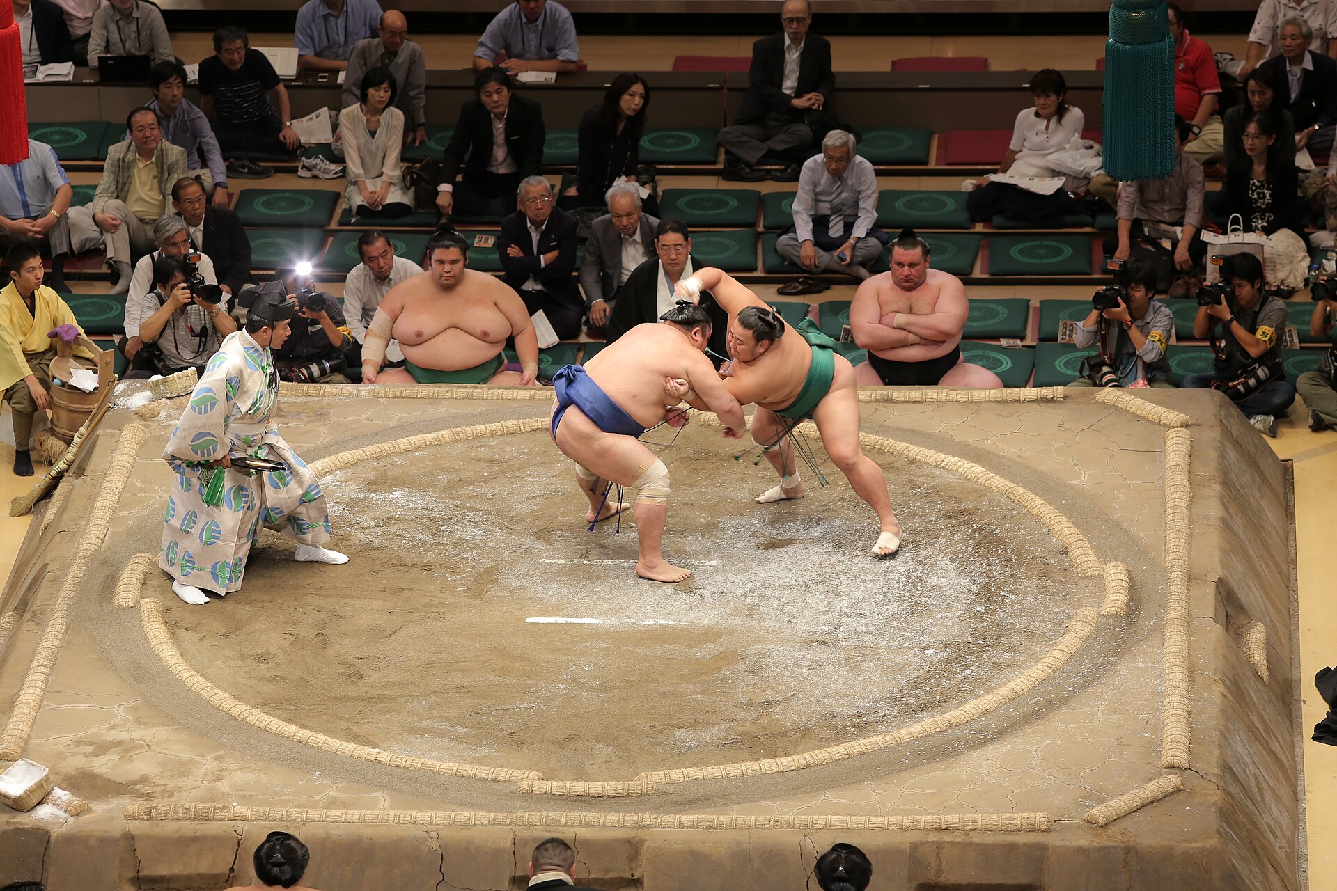 Overhead view of the clay sumo ring with two wrestlers engaged and a referee alongside.